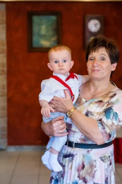 Reportaż z chrztu świętego Łódź babcia z wnukiem na rękach na tle ściany z zegarem 11